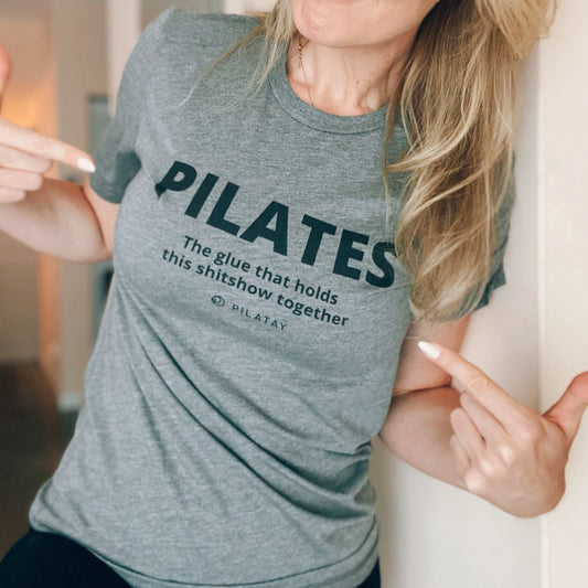 Grey t-shirt that reads Pilates the glue that holds this shitshow together worn by a Pilates teacher, pointing to the text.